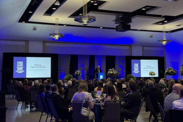 Mining 125th gala room with tables and a speaker at a podium with two screens displaying Mining Engineering Gala